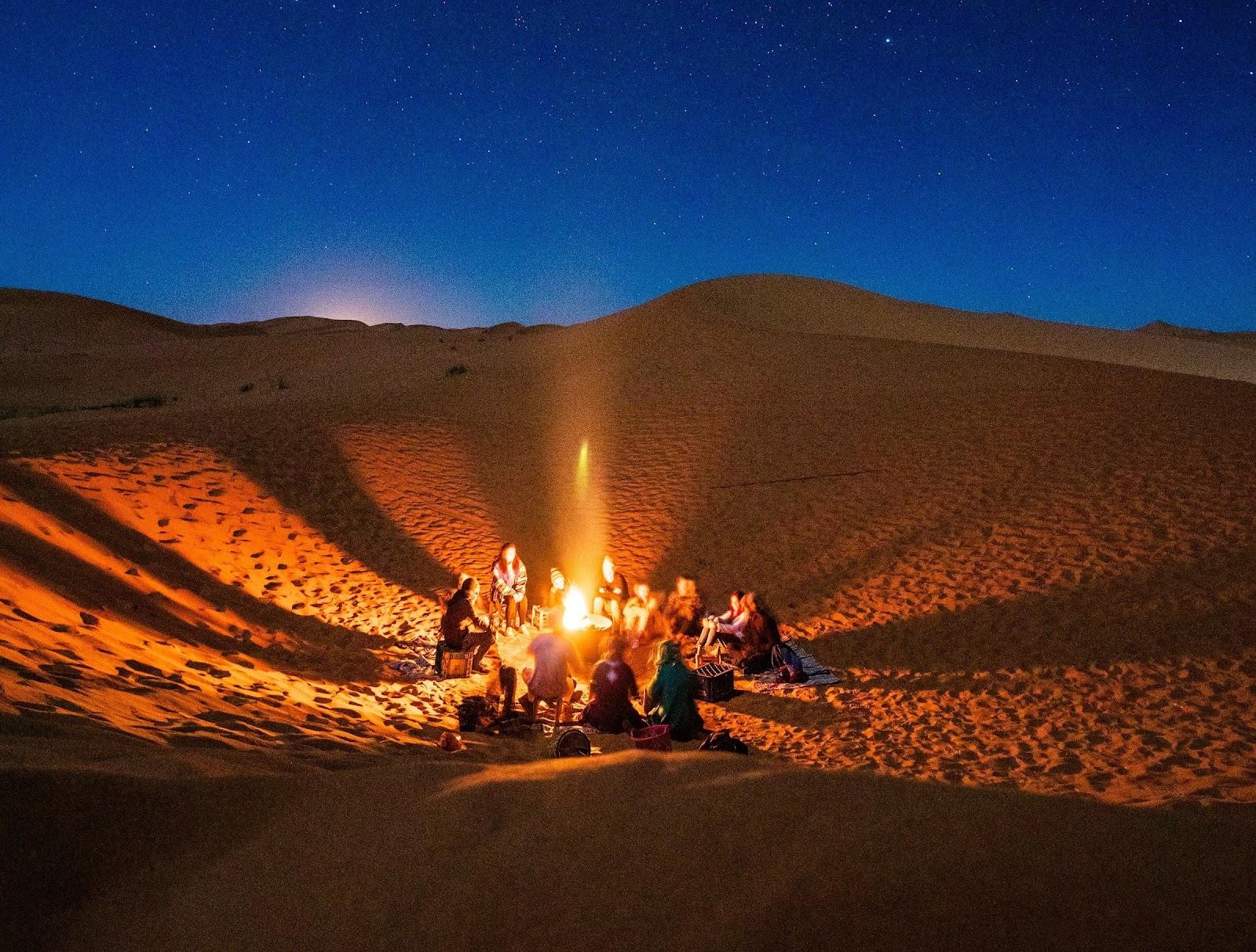 Tourists camping in the Sahara at desert at night, crowding round a fire.
