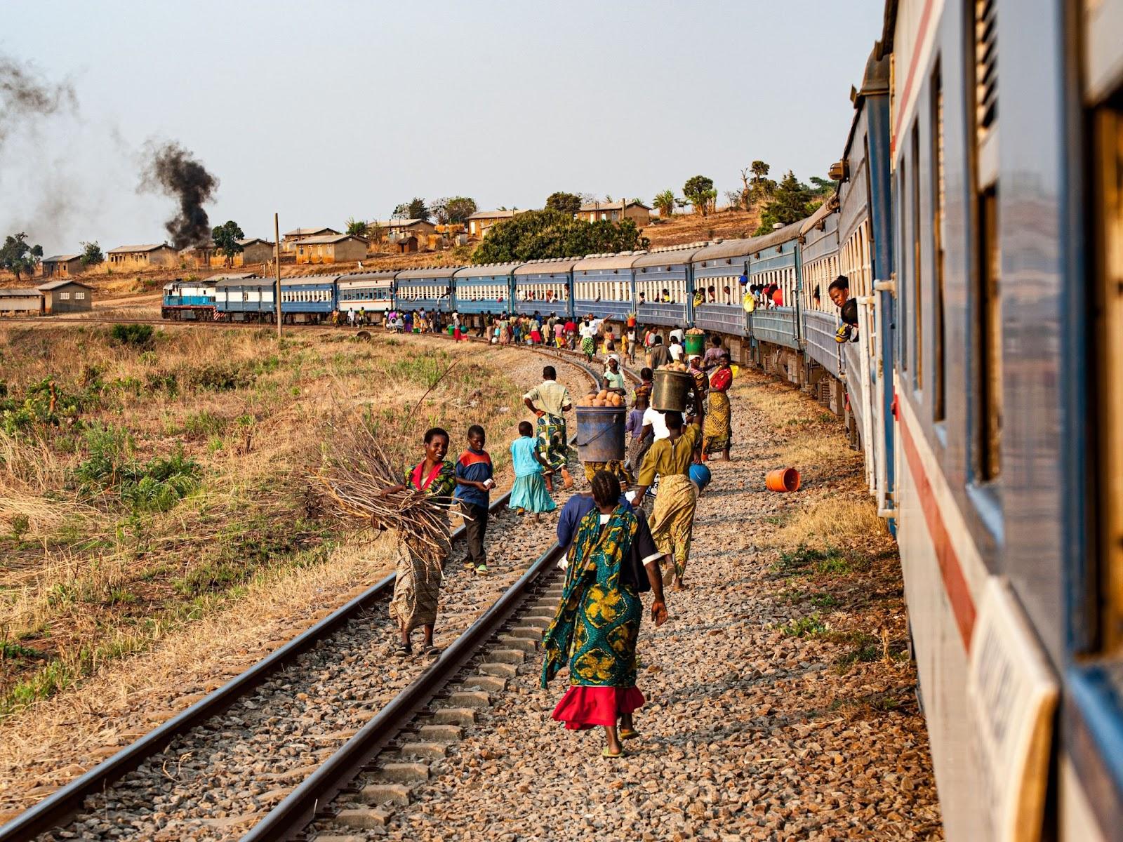 Train on the Tazara railway with lots of local people in brightly coloured, traditional clothes walking alongside