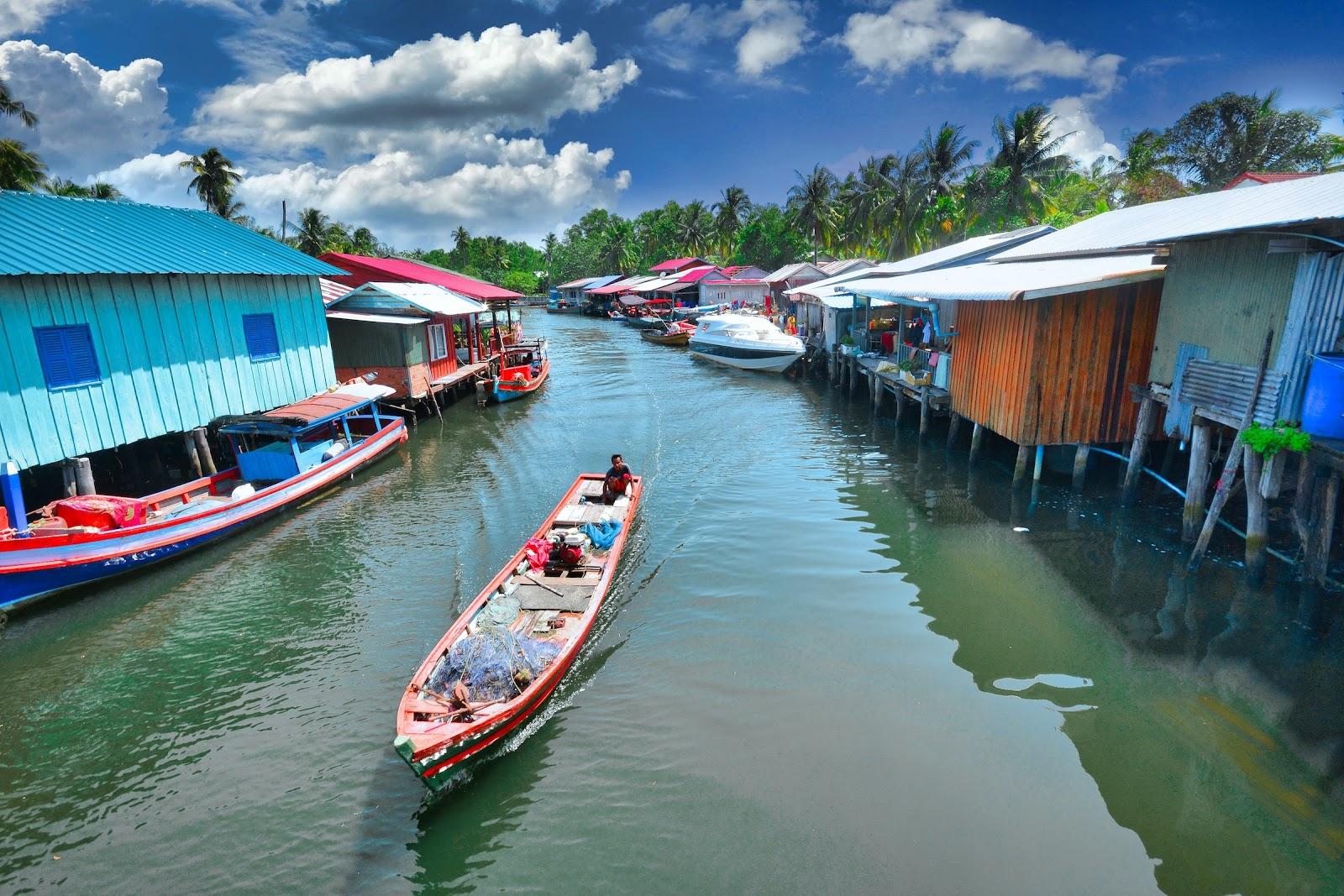 A colourful fishing boat on a narrow river with houses on stilts.