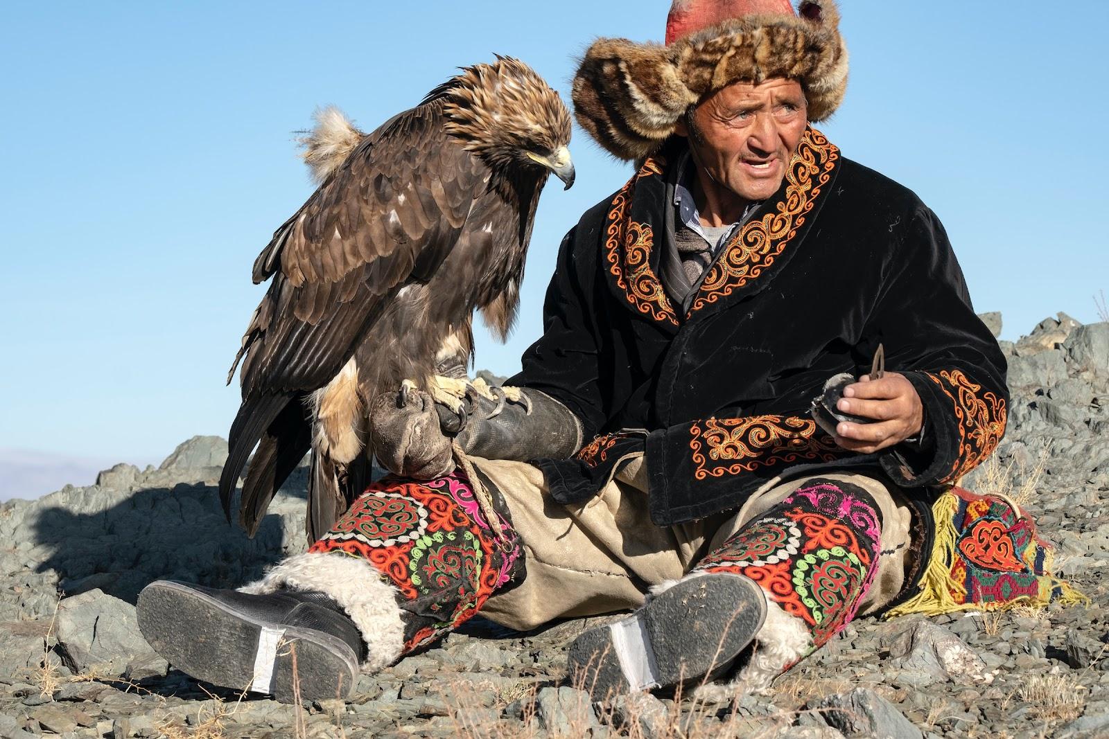 A Mongolian man sits with an eagle resting on his arm. 