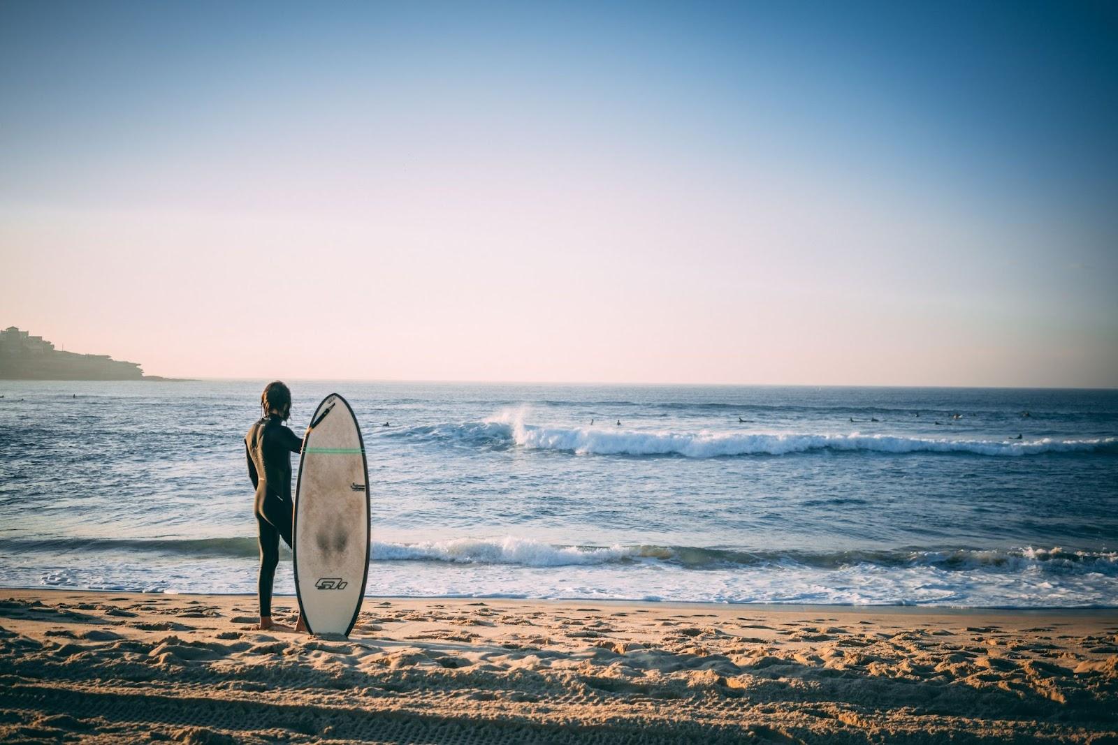 Surfer stood holding his surfboard on Australian beach looking out over the sea.