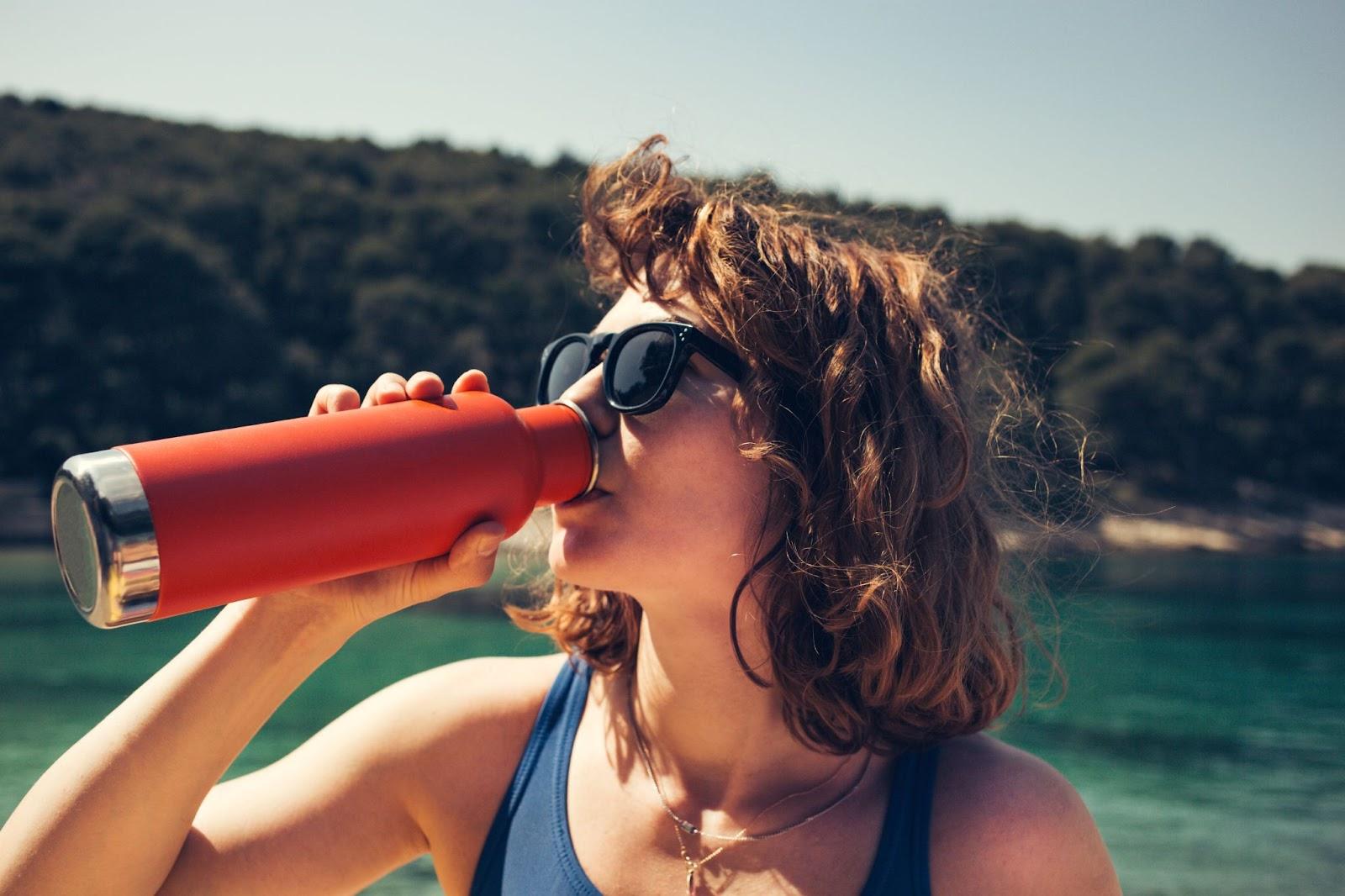A woman drinking from a reusable steel water bottle at the beach