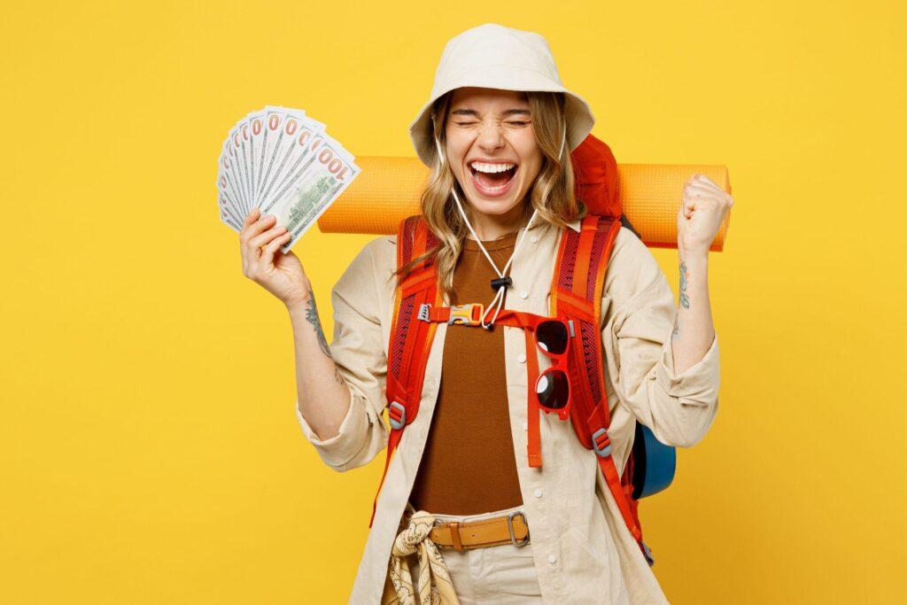 Young woman carring a travel backpack with a hand full of travel money. She is smiling. On plain yellow background.