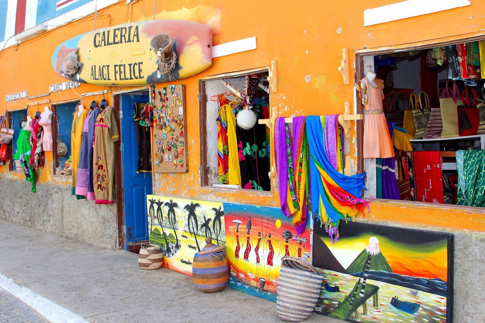 Sal. Cape Verde. Display of typical local products, paintings, crafts, clothing, souvenirs and surfboard on orange wall in outdoor street