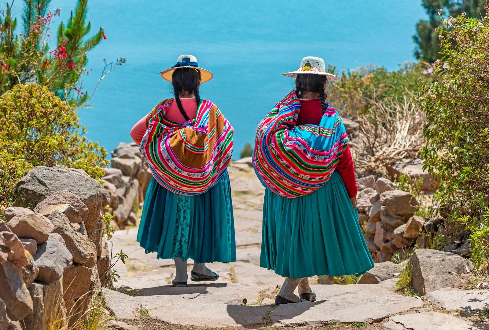 Two indigenous Quechua women in traditional clothes walking down the path to the harbor of Isla Taquile (Taquile Island) with the Titicaca Lake in the background, Peru