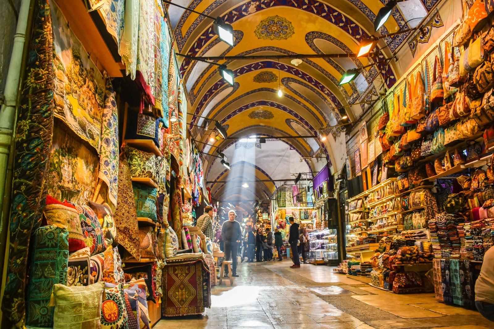 People shopping in the Grand Bazar, handmade pillows, bags and carpets are on the wall for sale. The sunlight comes to inside from roof window.