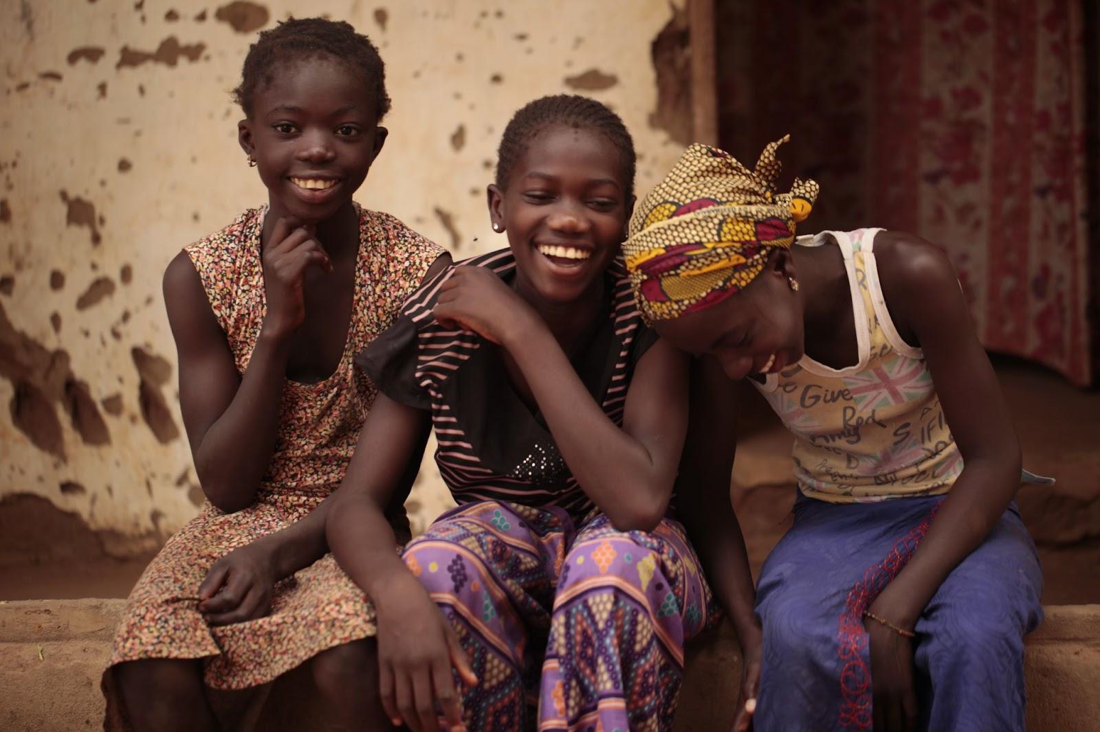 Buiba, the Gambia, Africa, girls sitting in front of an old white building with colourful material hanging in the door behind, outdoors on a sunny day