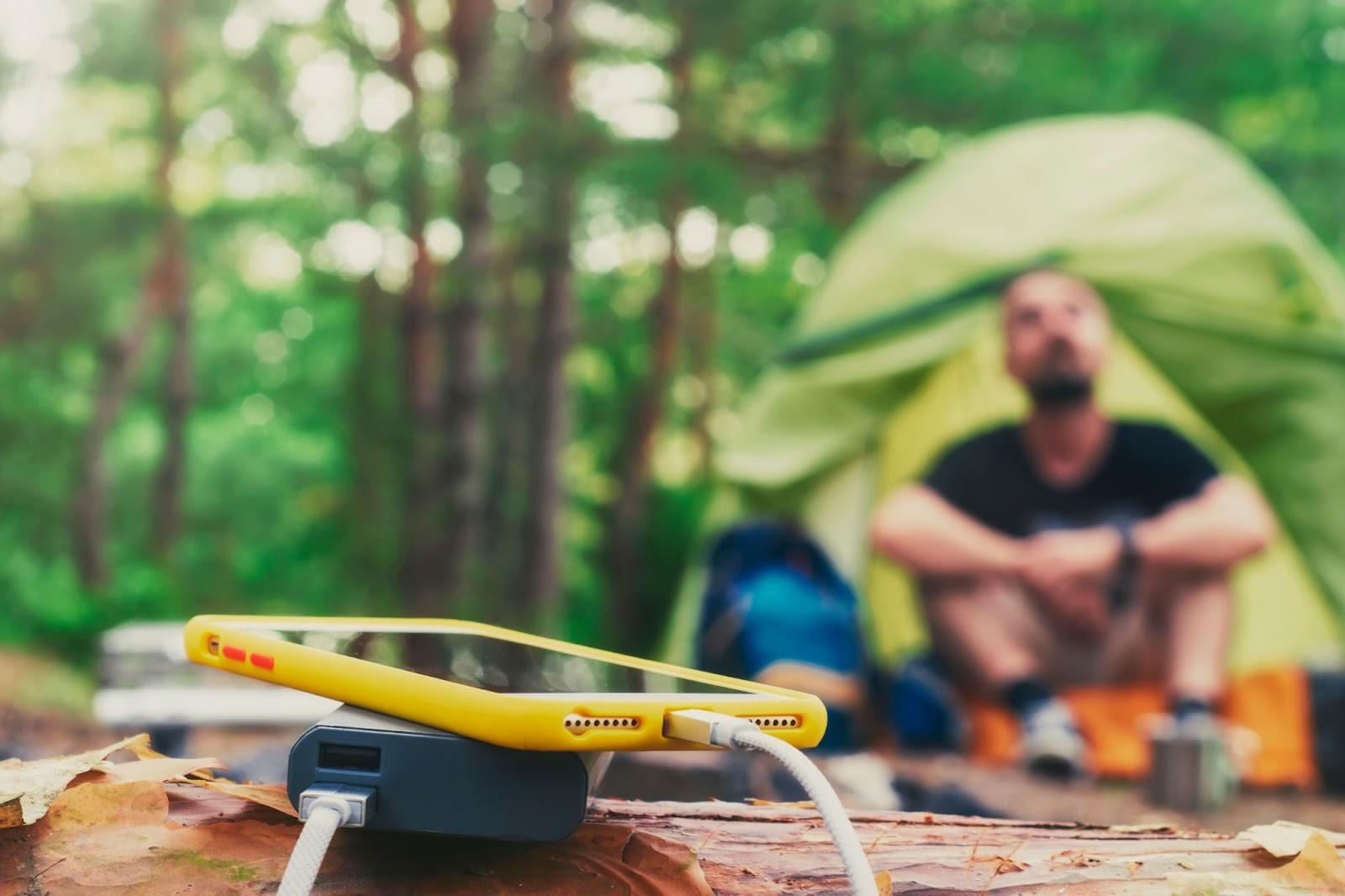 Smartphone is charged using a portable charger. Power Bank charges the phone outdoors with a backpack against the backdrop of a tent and a tourist