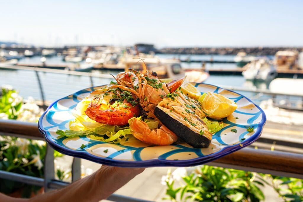 Serving Seafood and fish lunch in sea coast restaurant, Sicily, Italy. Italian food. Outdoor. Port with yacht on background