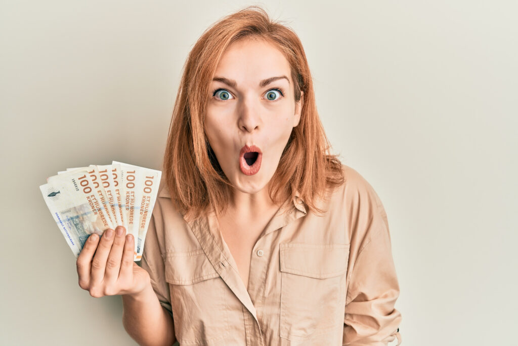 Young caucasian woman holding 100 danish krone banknotes scared and amazed with open mouth for surprise, disbelief face