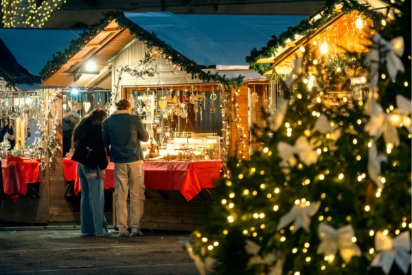 Unidentified people stand near illuminated wooden kiosk with souvenirs at traditional christmas market as christmas tree with lights in foreground in Asti, Italy