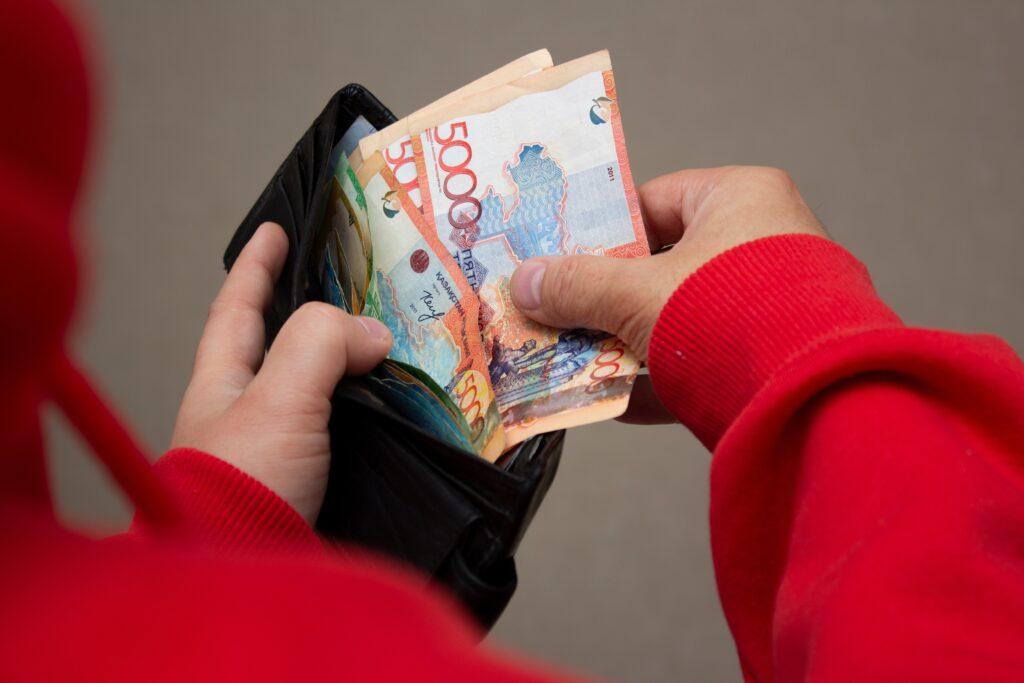 Man in red sweatshirt holds black wallet in hand with multicolored paper banknotes of Republic of Kazakhstan.