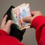 Man in red sweatshirt holds black wallet in hand with multicolored paper banknotes of Republic of Kazakhstan.