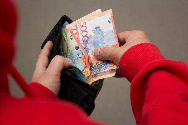 Man in red sweatshirt holds black wallet in hand with multicolored paper banknotes of Republic of Kazakhstan.