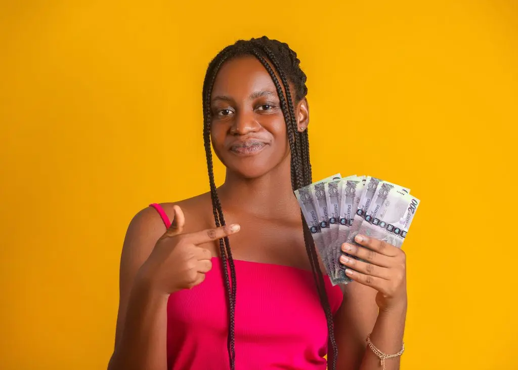 young woman with braided hair and a pink top, smiling as she holds a fan of Botswana Pula banknotes