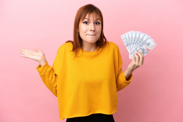 Redhead girl taking a lot of money isolated on pink background having doubts while raising hands